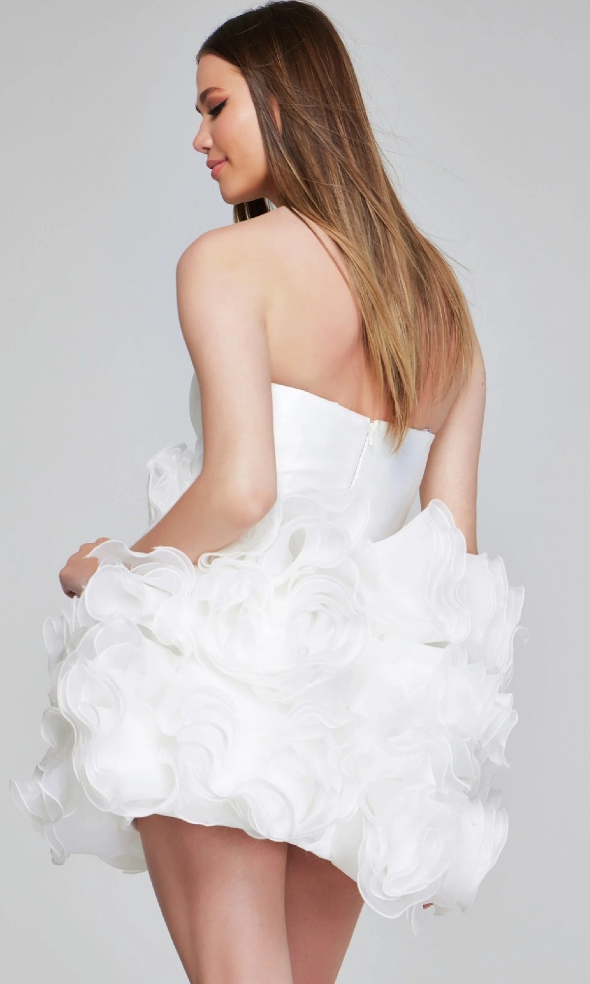 A woman wears the Strapless Short Rosette Hoco Dress: Jovani 40334, featuring a strapless design, voluminous ruffled skirt, and delicate 3-D rosettes, as she stands against a plain background.